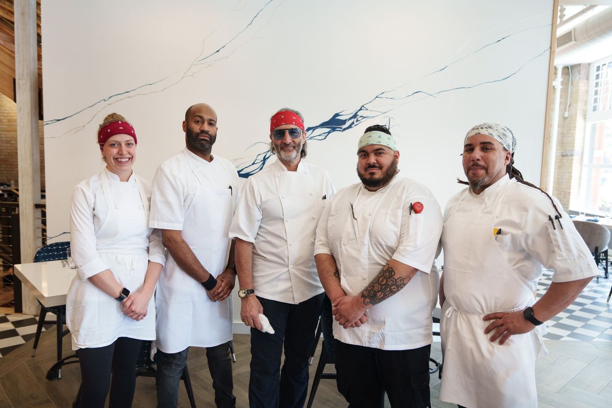 Five people wearing white chefs coats standing in a line with their arms folded in front of a white wall with a long blue streak running through it.