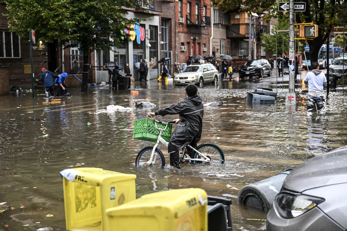A lone figure in a black raincoat appears to struggle to control a bike with a green basket on its front as they ride through water that covers the bottom half of the bike’s wheels. Partially submerged parked cars can be seen in the background, and flotsam bobs on the brackish, brown water.