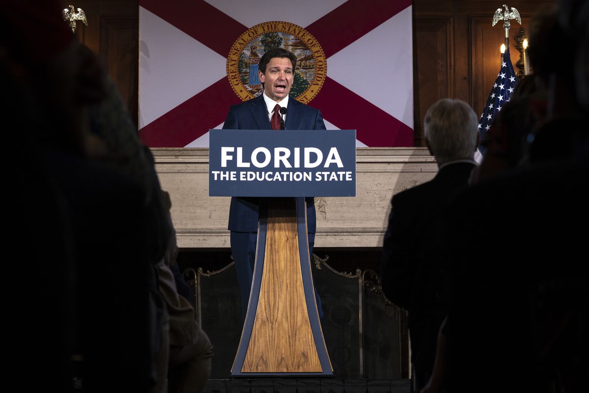 Ron DeSantis, wearing a navy suit and red tie, speaks from a wooden podium at the front of a crowded press room, with a banner saying “Florida: The Education State” in front of him.