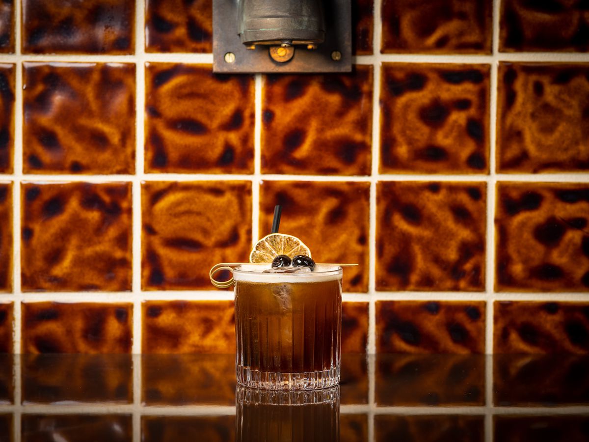 A dark cocktail with chocolate tones against a moody tile in a new bar.