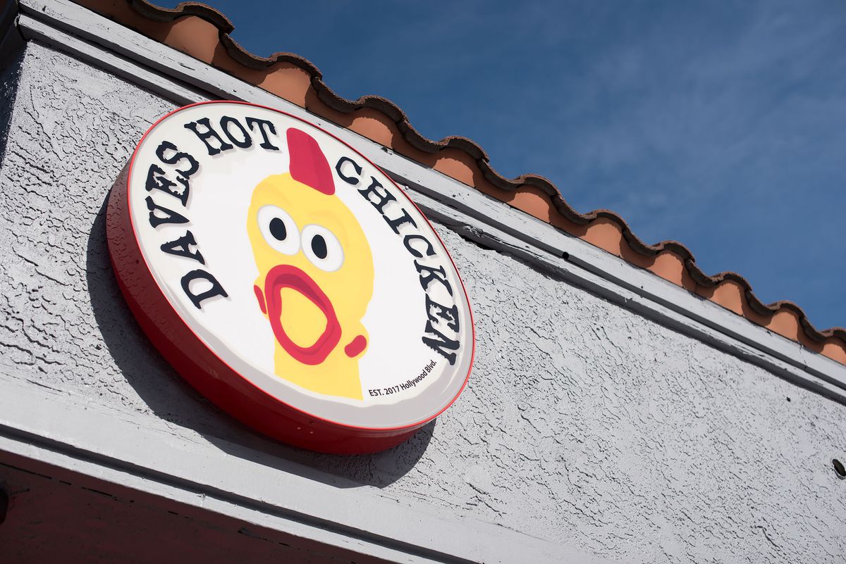 A circular sign with a chicken and the words “Daves Hot Chicken” hangs from a building.