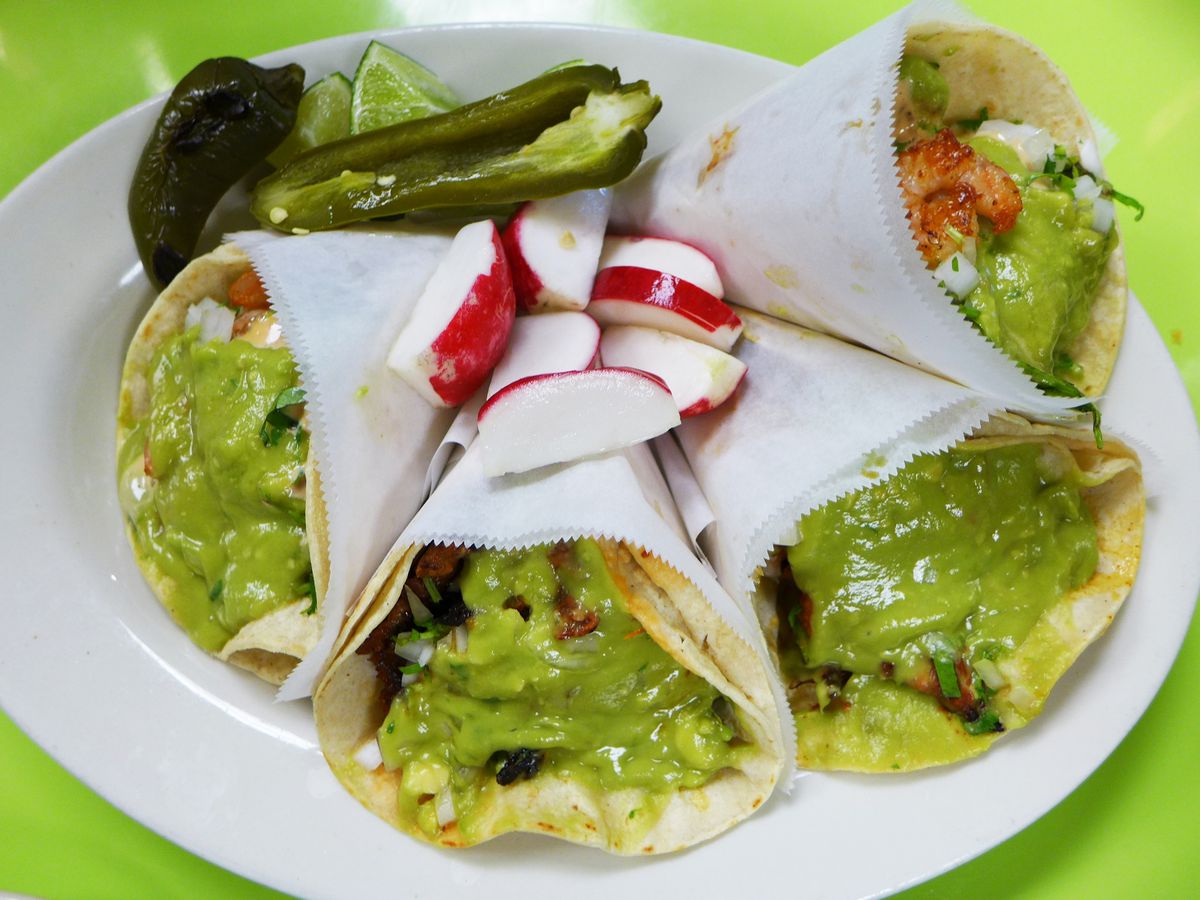 Three cone shaped tacos with guacamole spilling out and chiles and radishes on the side.