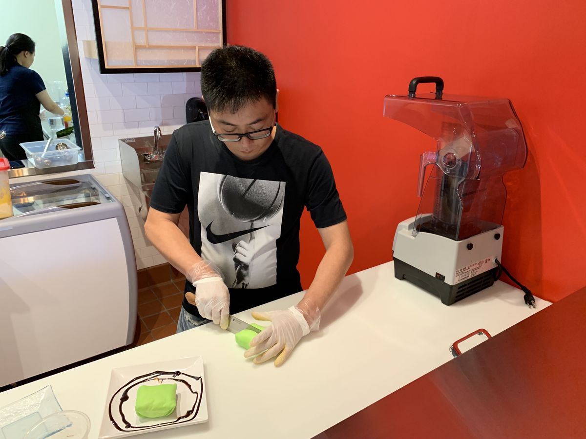 Billy Ho slices durian pancakes on a kitchen counter with a blender on the side of the counter and a red wall in the background.