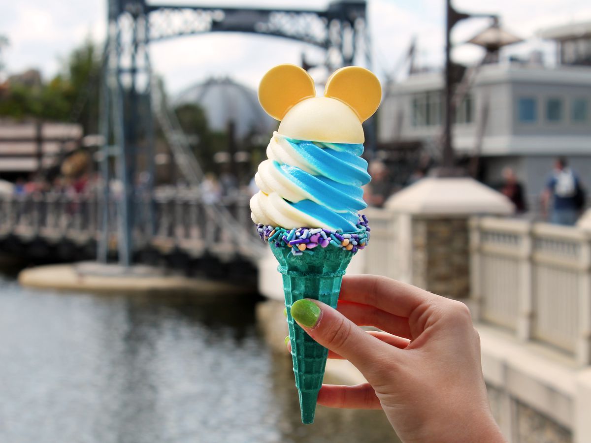 A teal-colored ice cream cone of white and blue soft serve swirled with a rim of sprinkles around the cone and cookies in the shape of Mickey Mouse ears on top