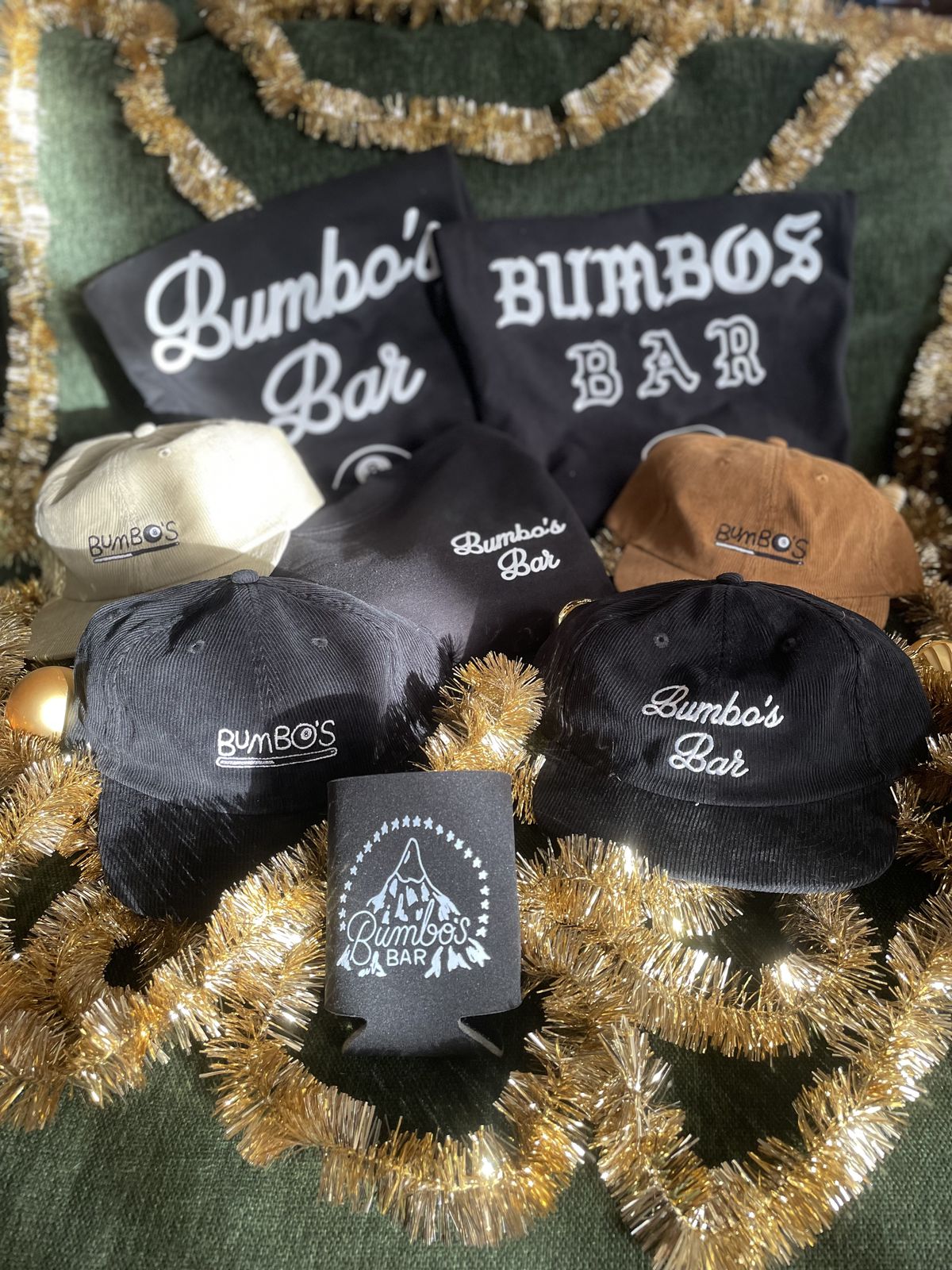 Baseball hats in black, brown, and tan, with Bumbo’s Bar embroidered, a beer cozy, with gold garland.