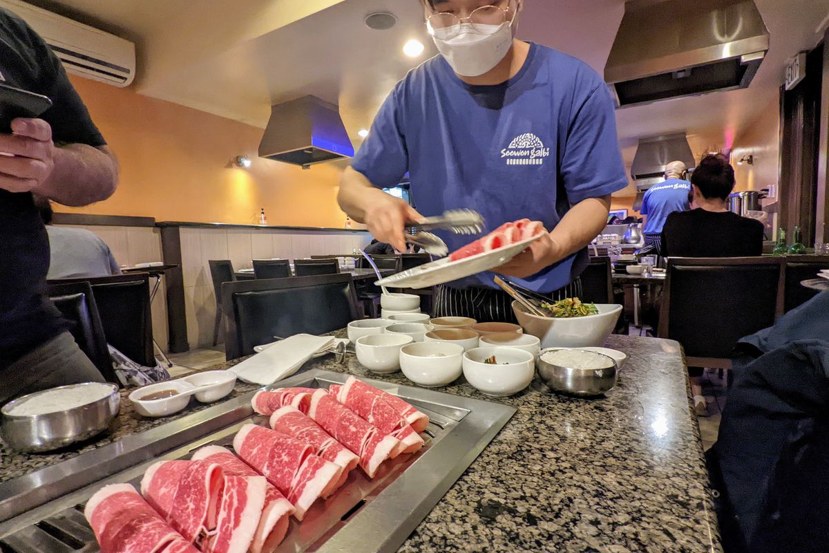 Blue-shirted server wearing a mask places raw beef slices on a tabletop grill.