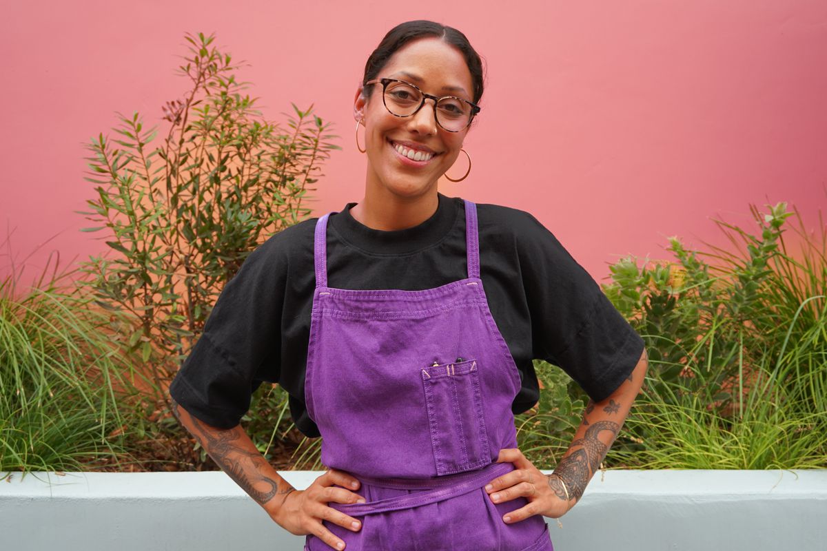 A woman in glasses and a purple apron smiles at the camera.