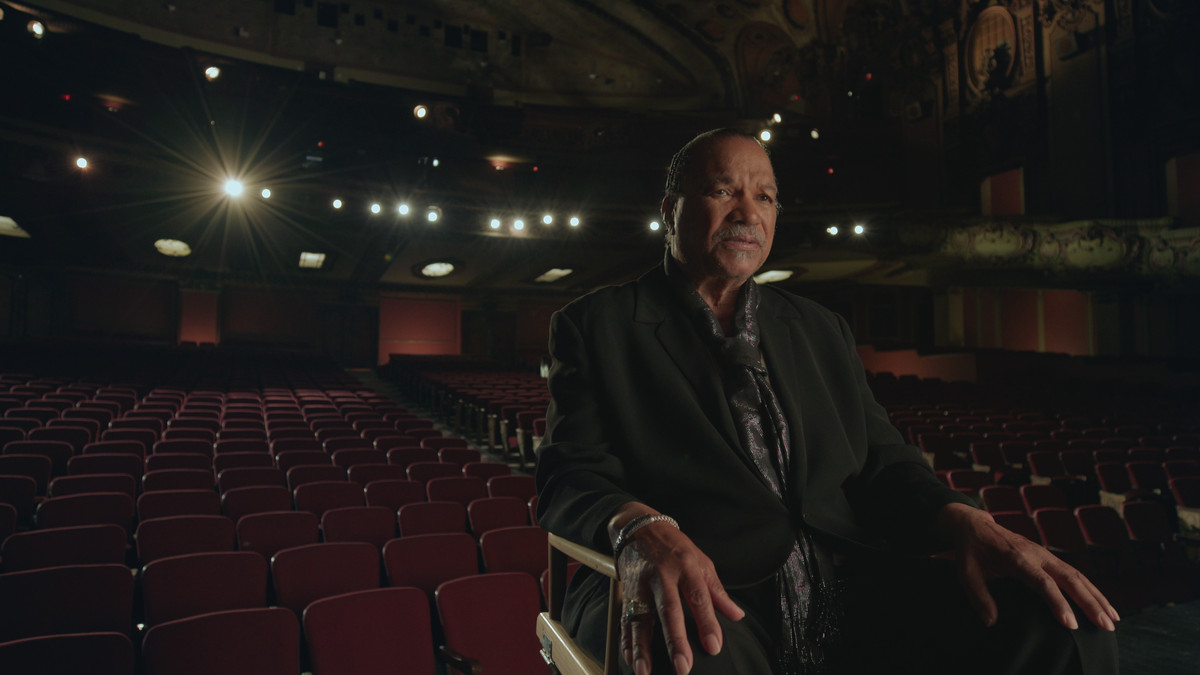Billy Dee Williams se sienta en la silla de un director en el escenario de un teatro, de espaldas a las sillas vacías del público