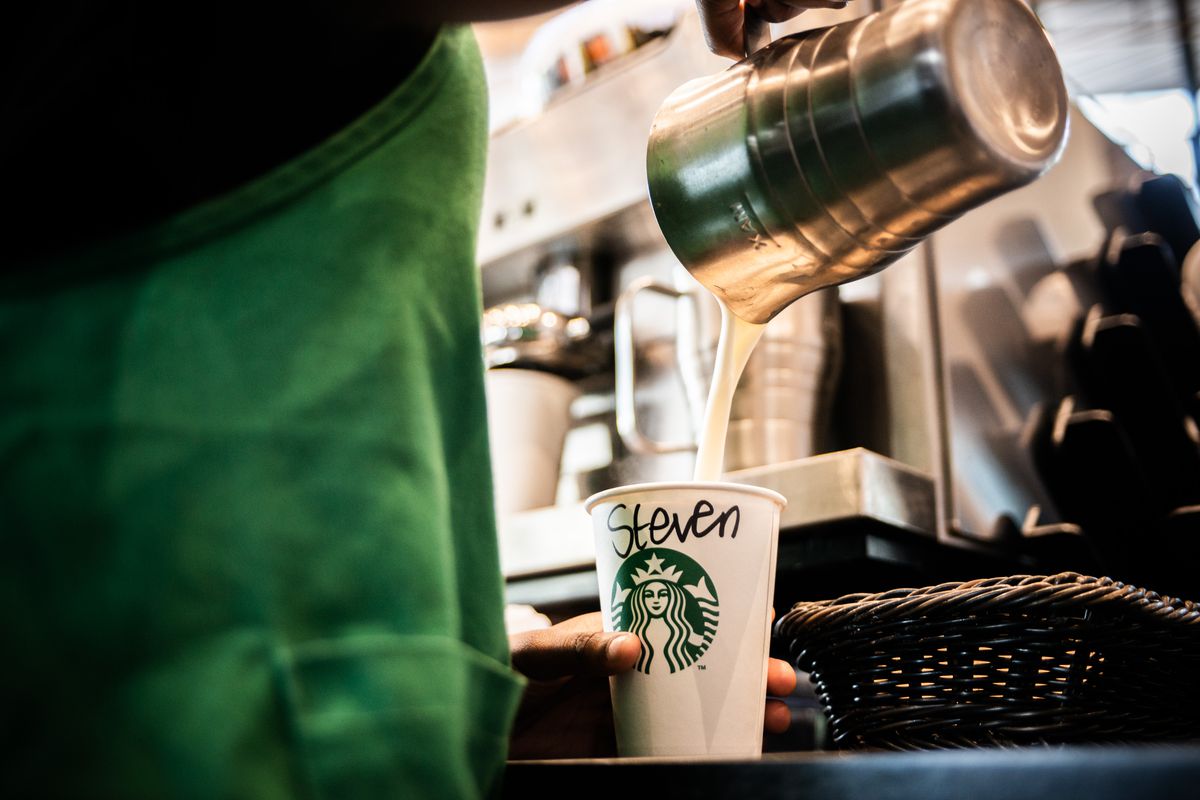 A person in a green apron pours milk from a pitcher into a white Starbucks to-go cup.