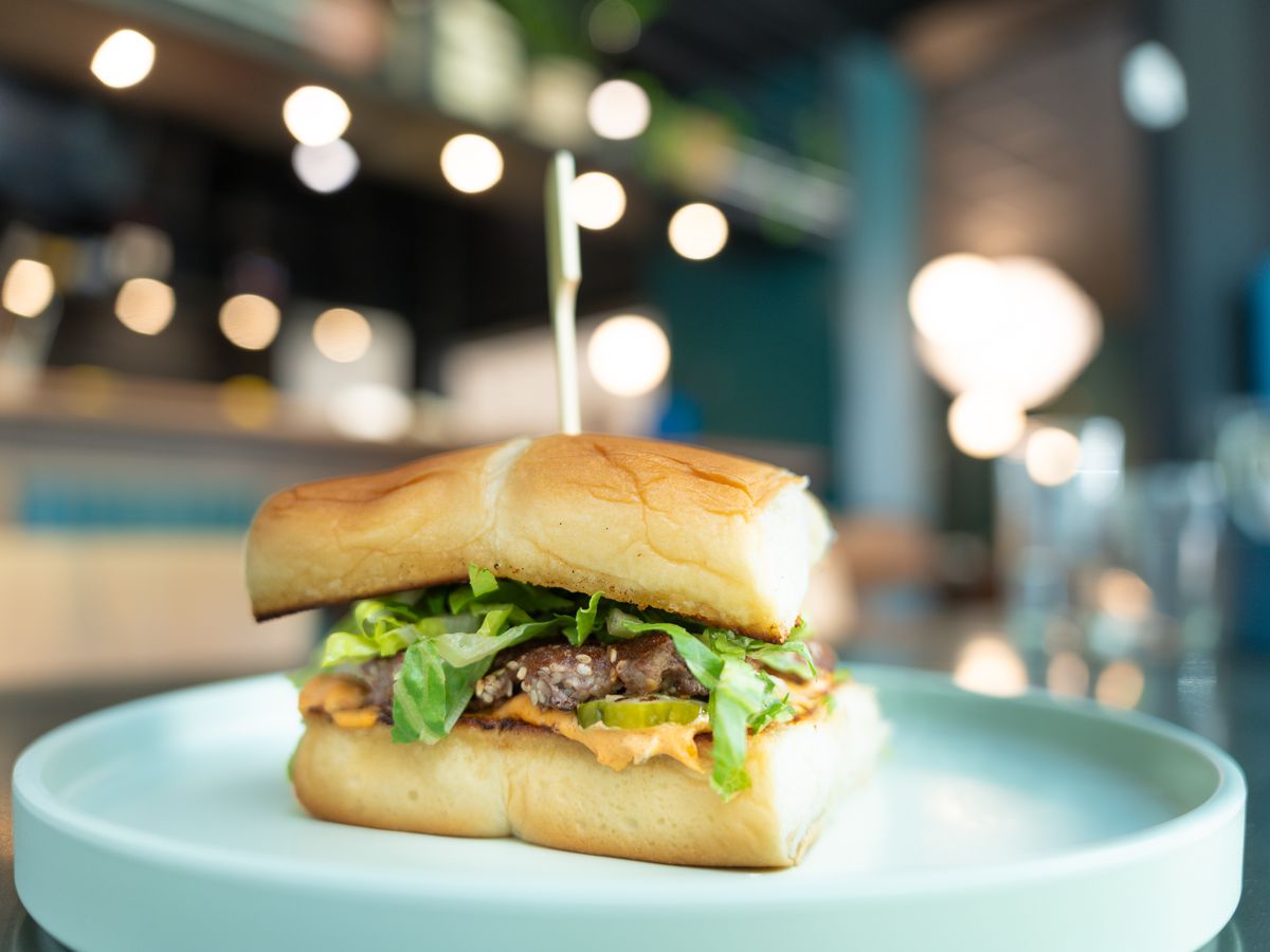 A burger with lettuce and cheese on a square of four Hawaiian rolls, on a white plate, with a restaurant’s interior visible but blurred in the background.