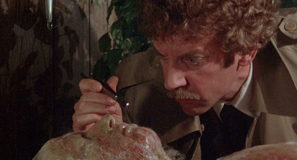 A man (Donald Sutherland) examines the face of a body trapped in a strange sinusoidal skin resembling a spider's web.