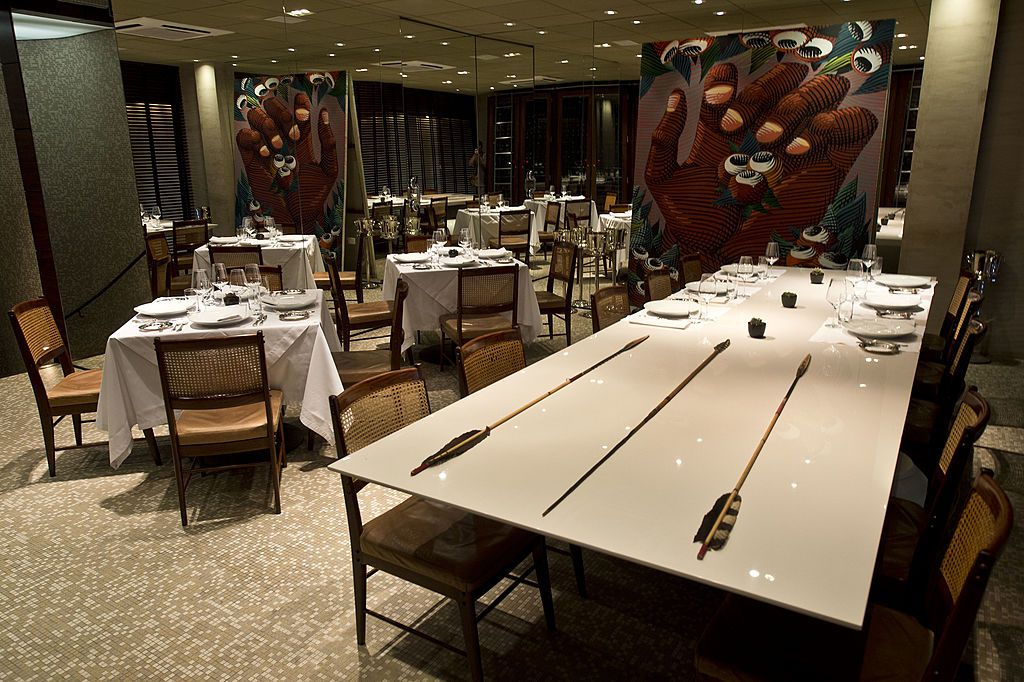 An empty dining room decorated with massive pictures of someone’s hand holding tiny dishes, white table cloths and place settings on tables, and a large communal table in the foreground painted with long arrows