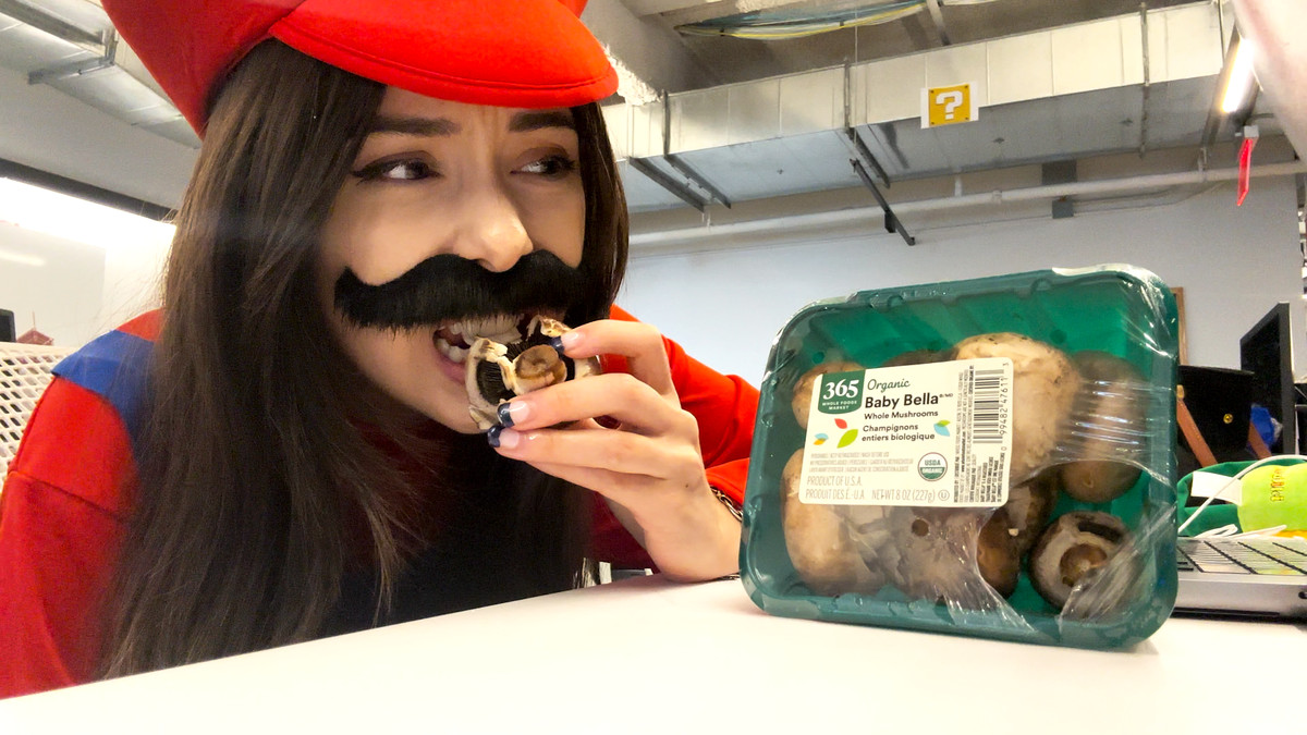 Tina, dressed as Mario, grimacing as they bite into a raw Baby Bella mushroom.