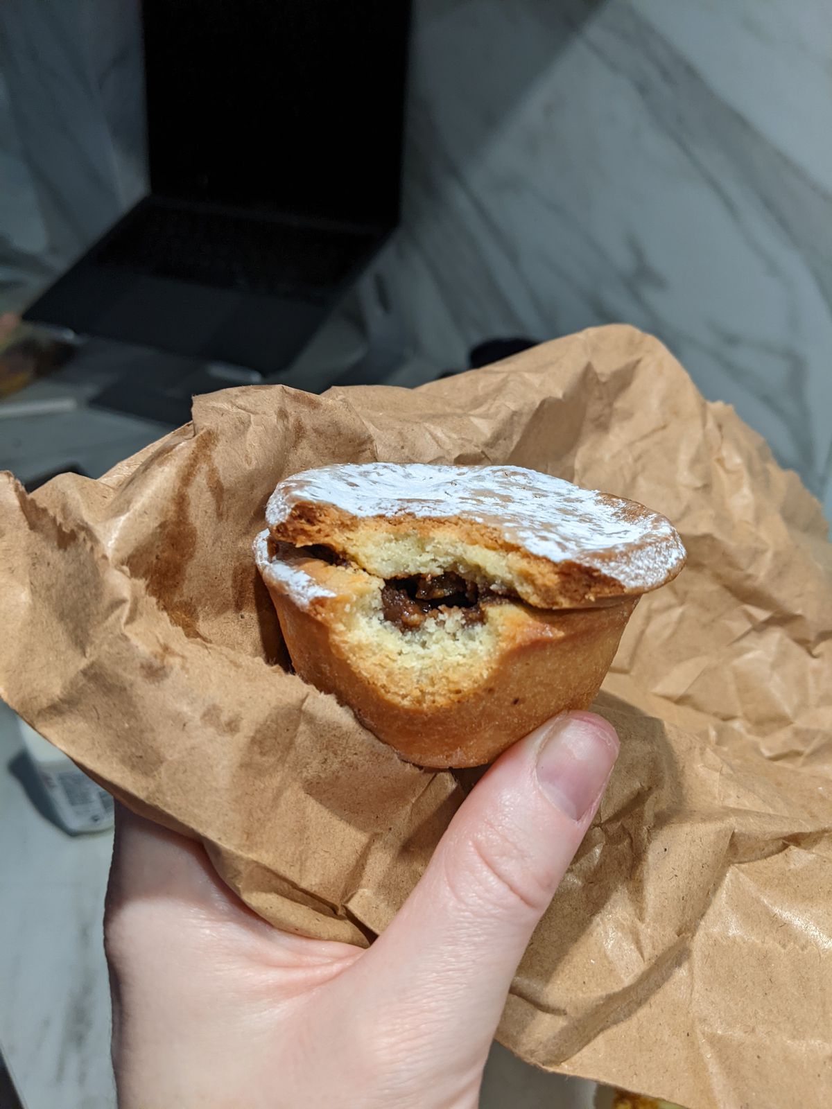 A hand holding a bitten-into mince pie, the filling visible, on top of a brown paper bag.