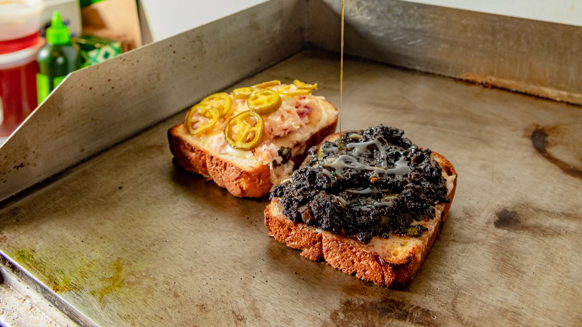 A hand drizzles hot honey out of a squeeze bottle onto an open-faced sandwich heating up on a flat-top grill.