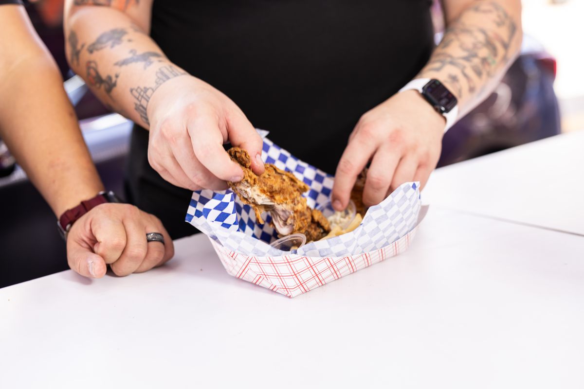 A man breaks apart a piece of deep fried turkey that’s in a cardboard boat with blue checked paper. Another man’s right hand sits in the frame.