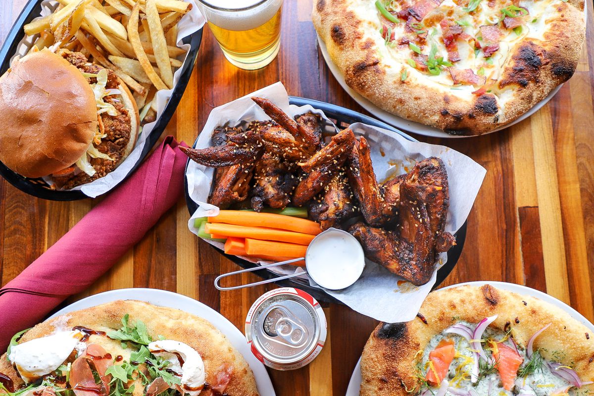 A table packed with pizzas, burgers, sandwiches, and beers.