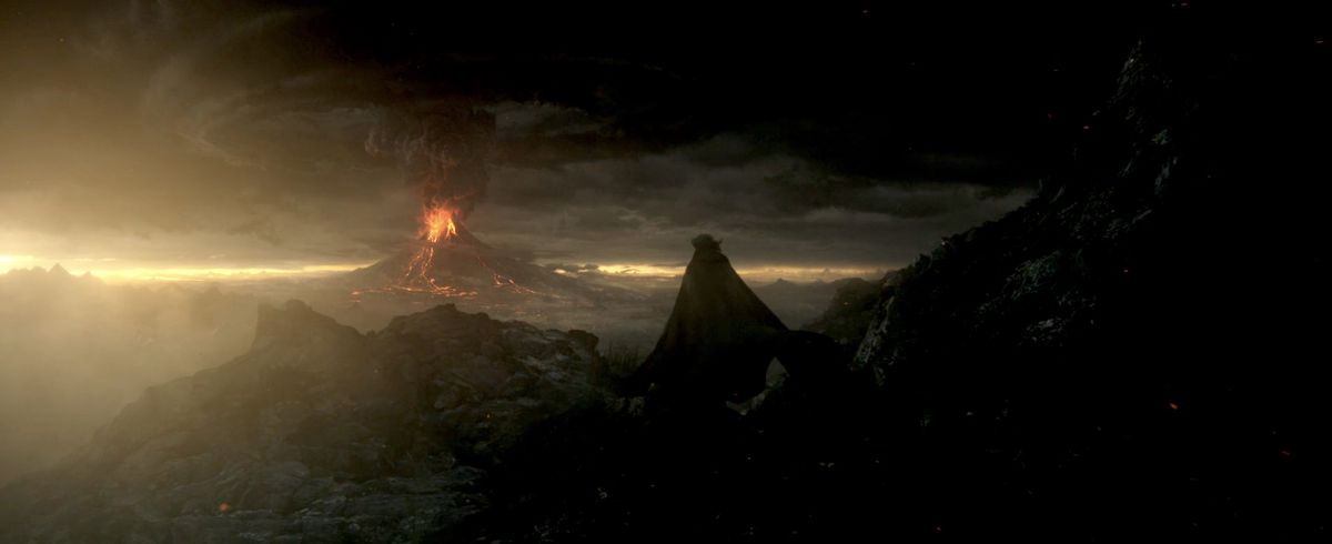 Sauron in his black cloak ascends a cliff to see Mount Doom firing off some lava and black smoke which clouds the rising sunset in The Rings of Power