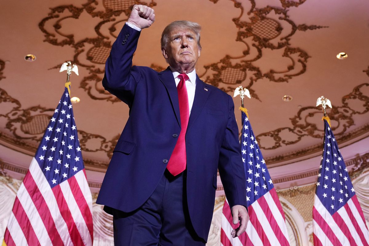 Former President Donald Trump wears a navy suit and red tie, stands in front of three American flags while raising closed fist in the air. 
