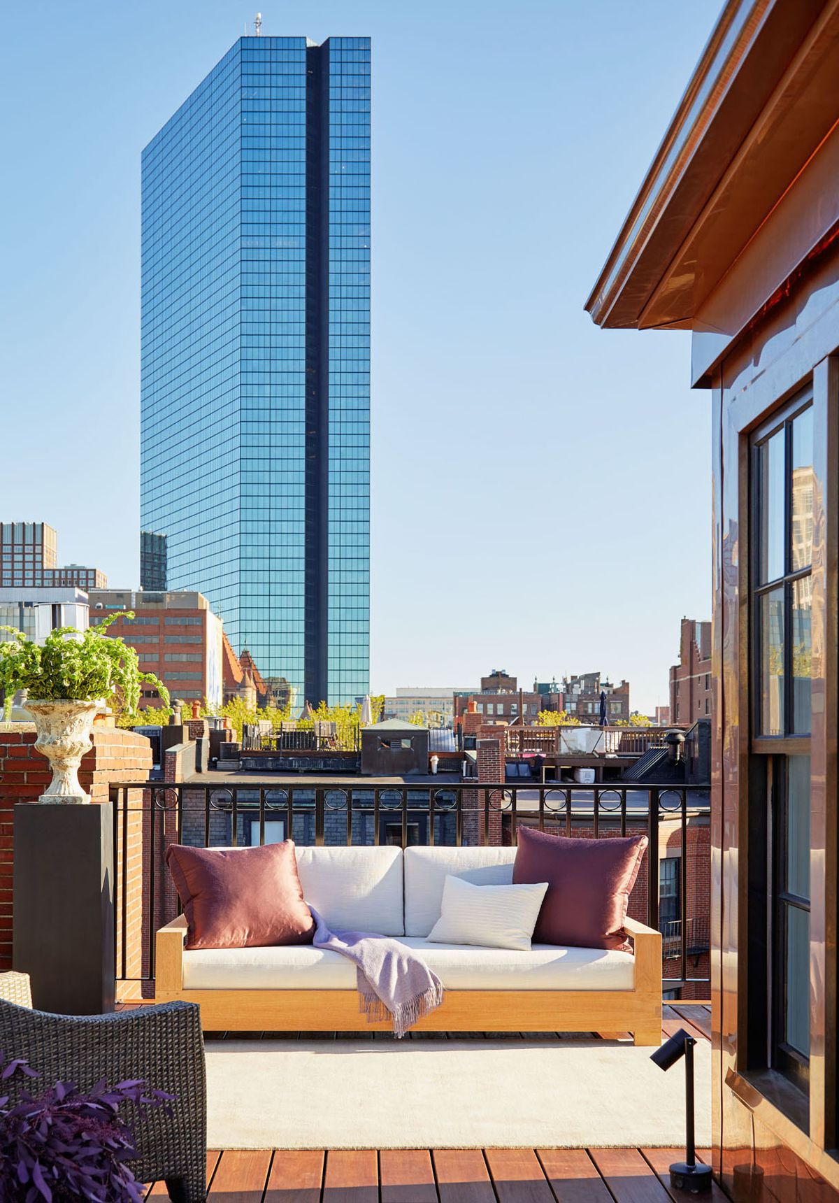 A sun-drenched view of a modern, outdoor loveseat with throw pillows on a private roof deck. A large shiny skyscraper is visible in the background.