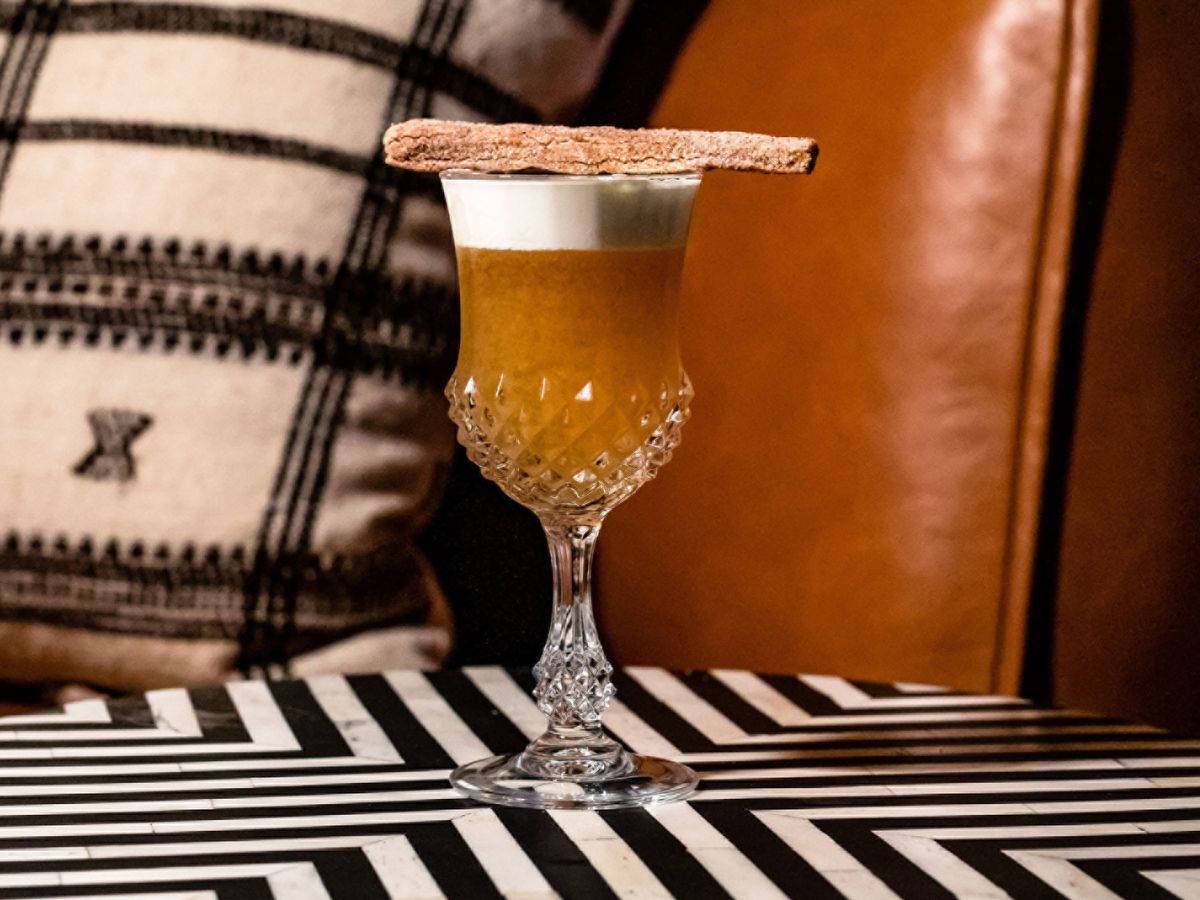 A brown cocktail in a goblet topped with a bread garnish.