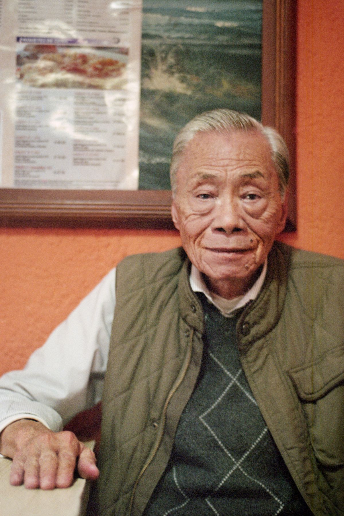 An elderly Chinese man smiles while sitting at a table at his restaurant.