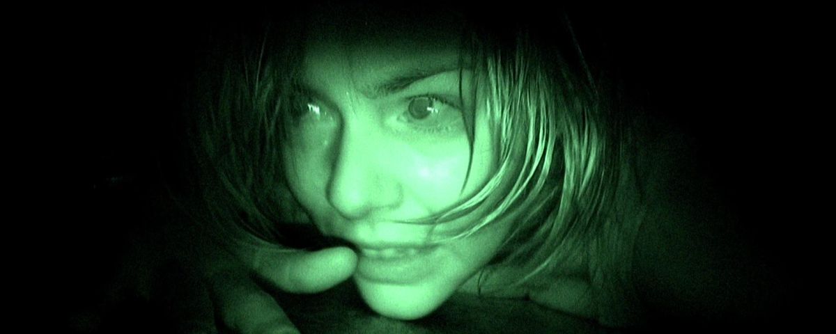 A grainy extreme close-up of the face of a frightened woman crawling along the floor, green-tinted as if seen through a night-vision camera in 2007’s [REC]