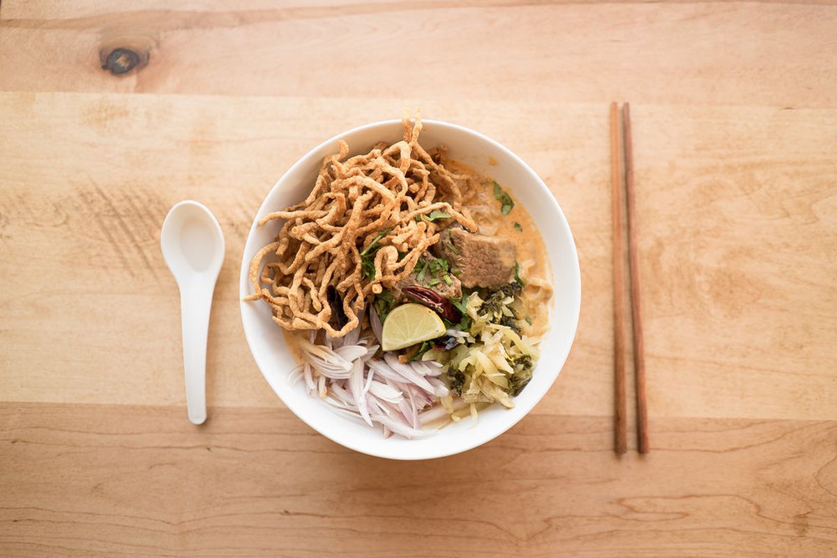 Overhead view of a white bowl full of a yellow curry with crispy noodles, preserved greens, a lime wedge, and red onion slices. It sits on a light wooden table with a spoon and chopsticks next to it.