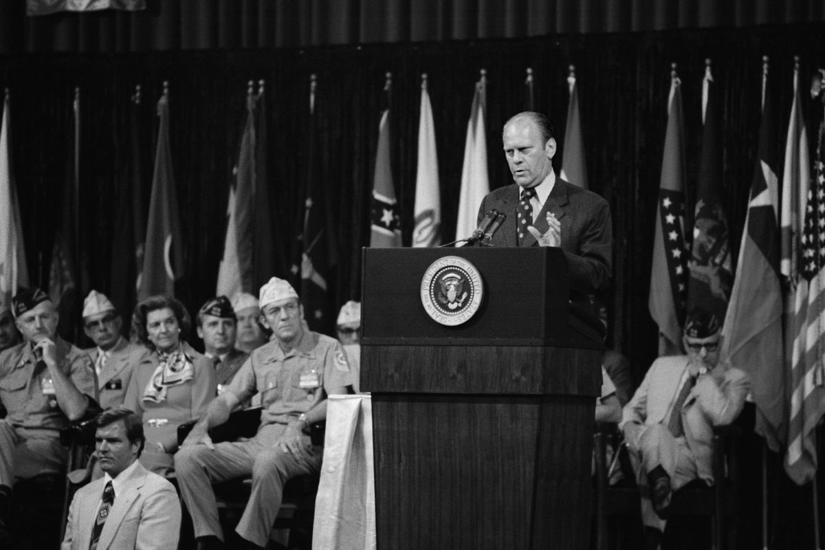 President Gerald Ford speaking onstage at a podium with uniformed service members seated behind him, circa 1974.