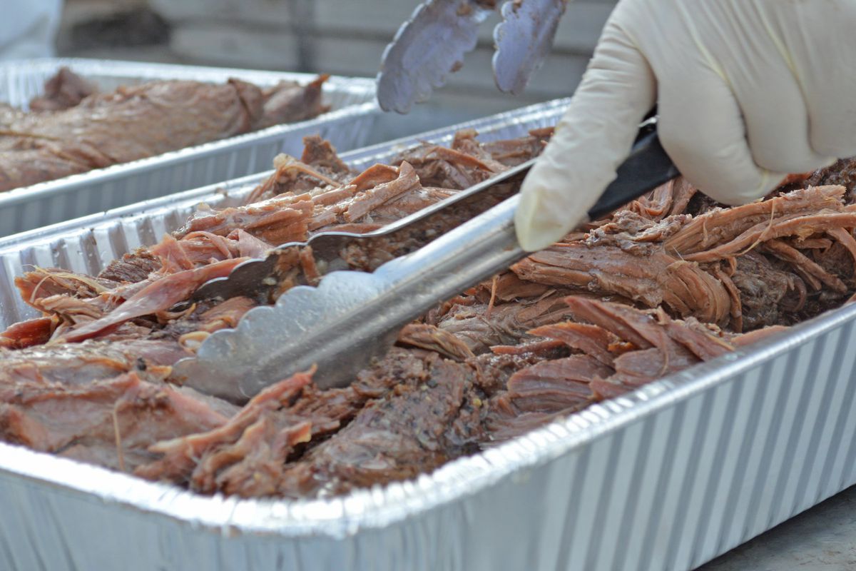 A gloved hand uses tongs to move around a large tray of pulled meat.
