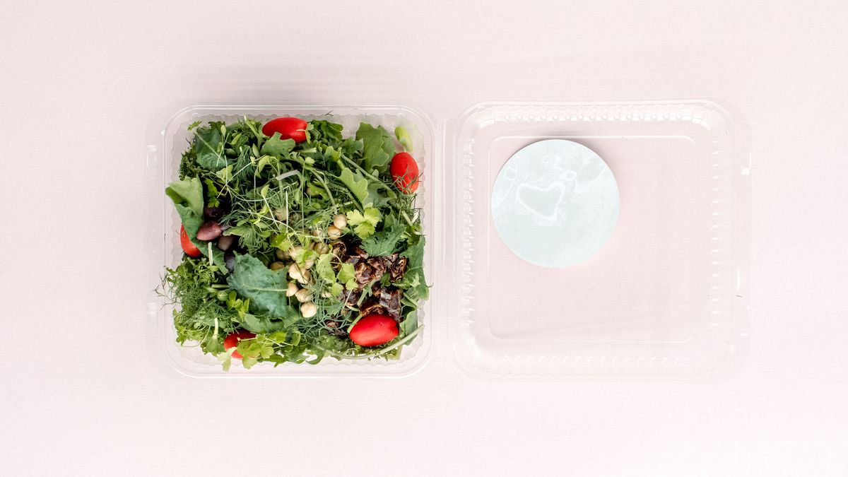 The Moroccan salad from Planted Detroit sits in a plastic container on a light pink background. It has tomatoes, greens, olives, and chickpeas.