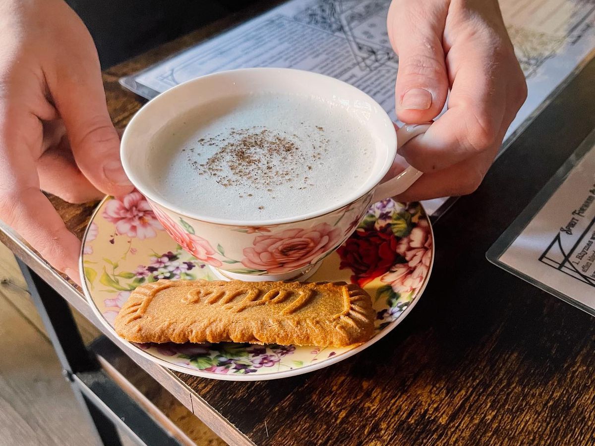 A person delivers a creamy cocktail served in an ornate tea cup, on a small rainbow platter with a cookie