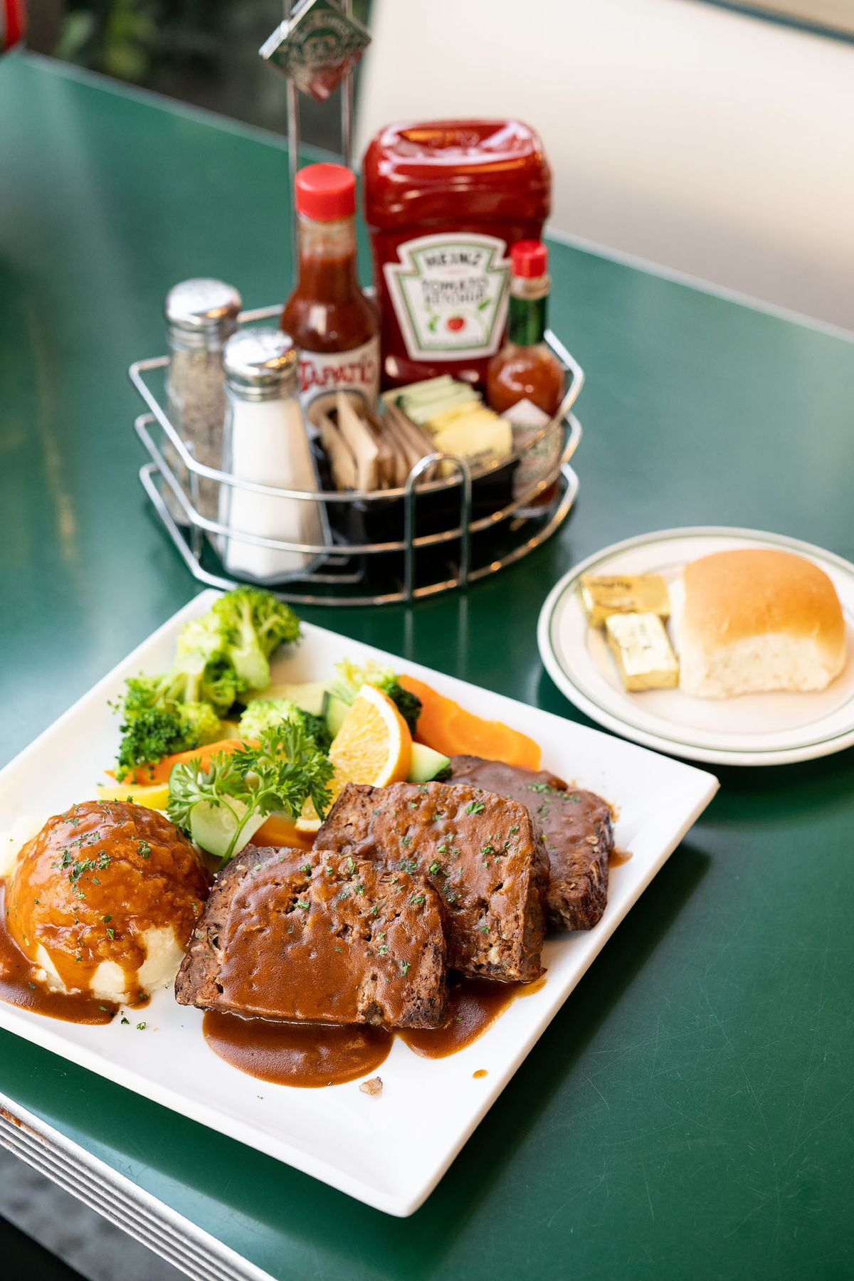 A green Formica table with saucy slices of meatloaf near a caddy of condiments.