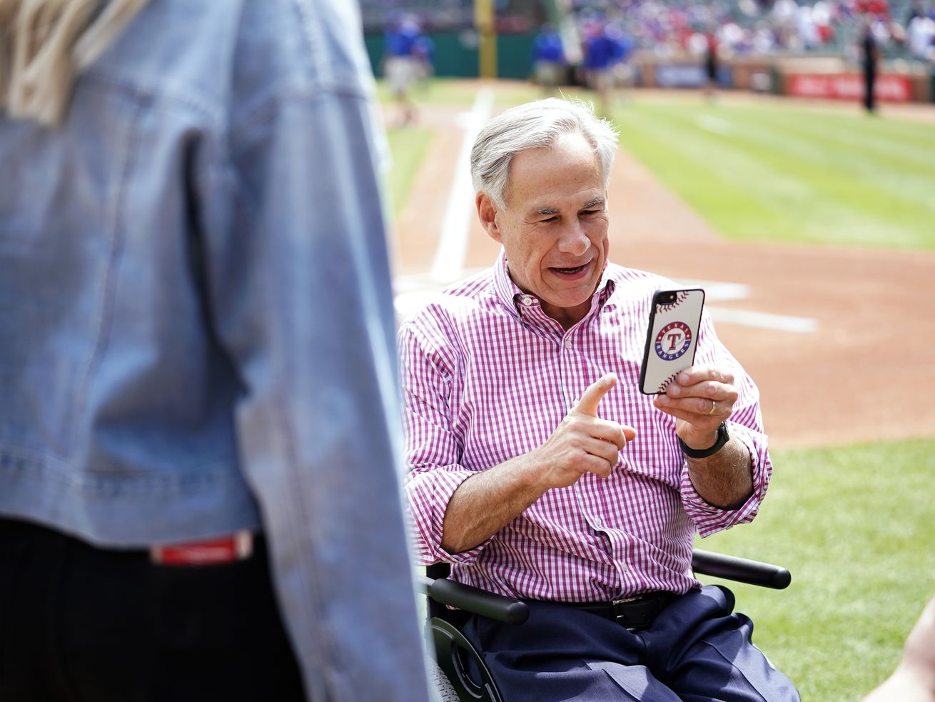Texas Governor Greg Abbott looks at his phone while seated on a baseball field.