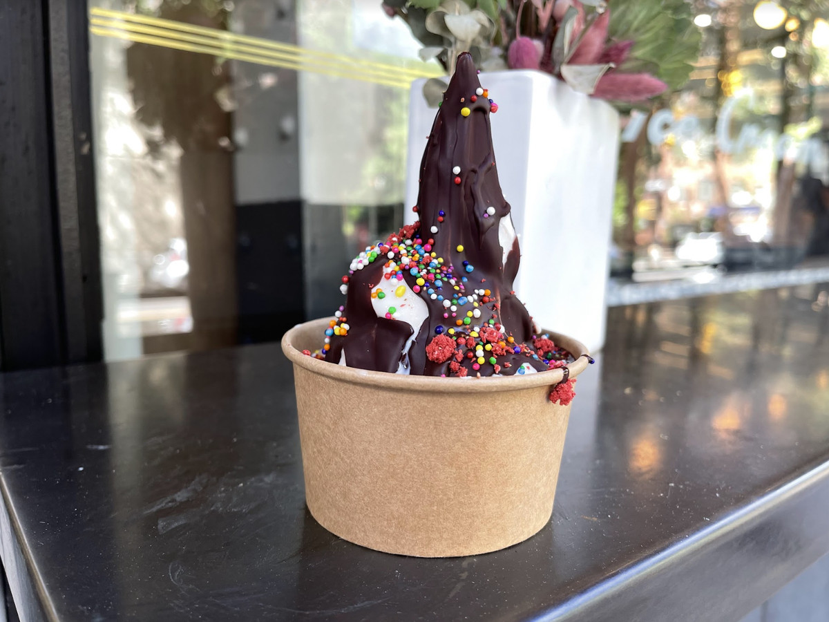 Soft serve in a brown paper cup is topping with sprinkles and chocolate sauce.