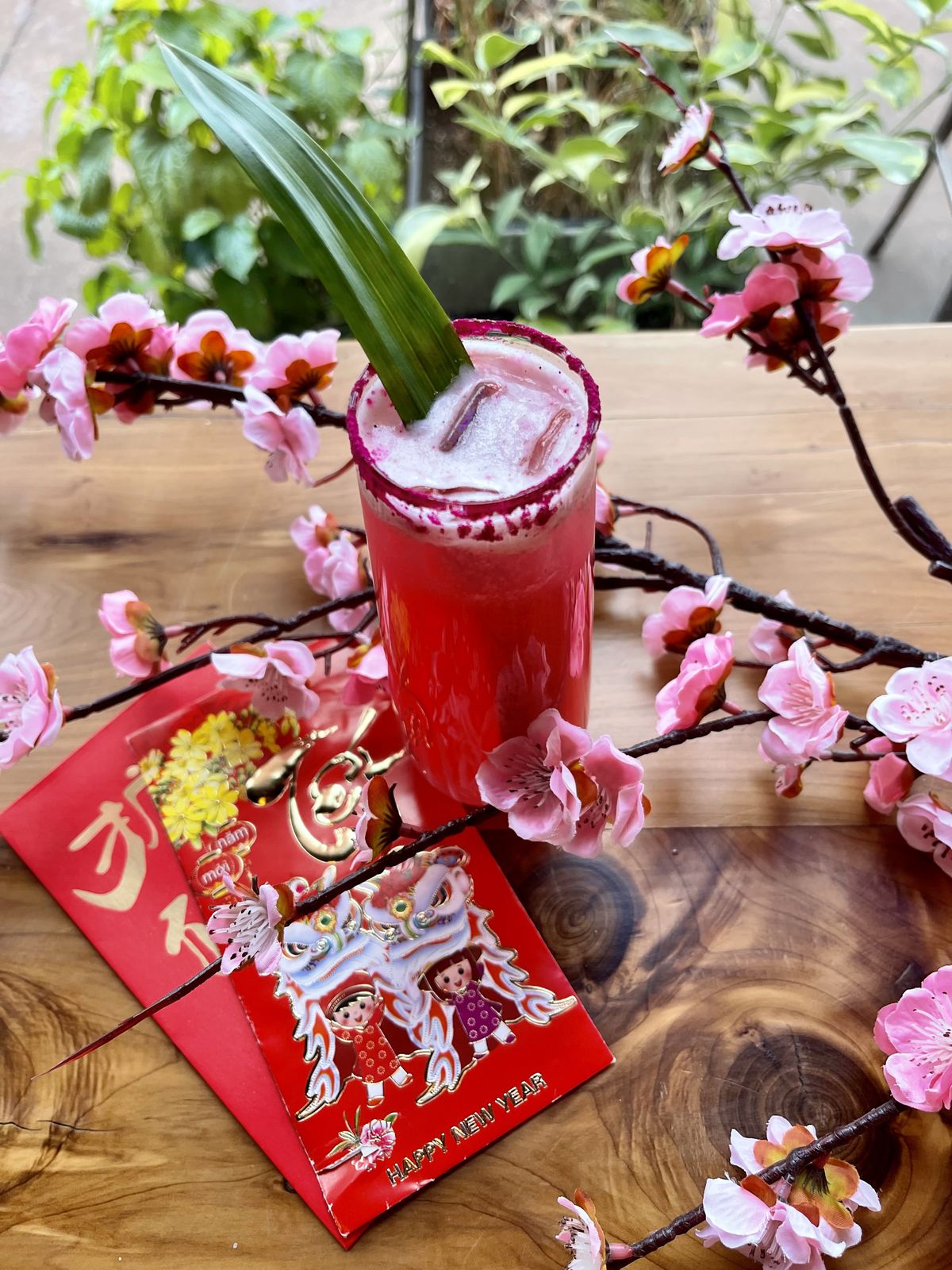 A pink cocktail sits on a wooden table with cherry blossoms around it. 