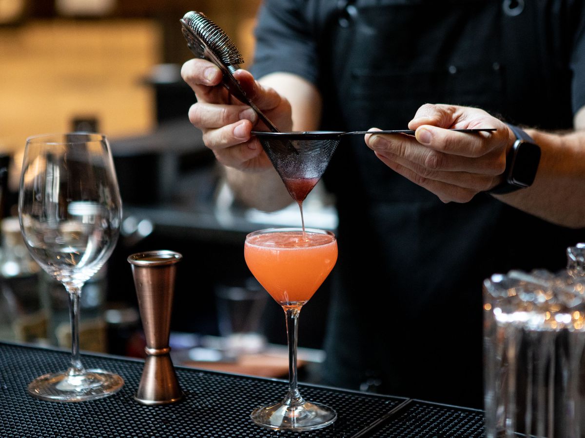 A bartender strains liquid through a sieve into a glass full of pink liquid.