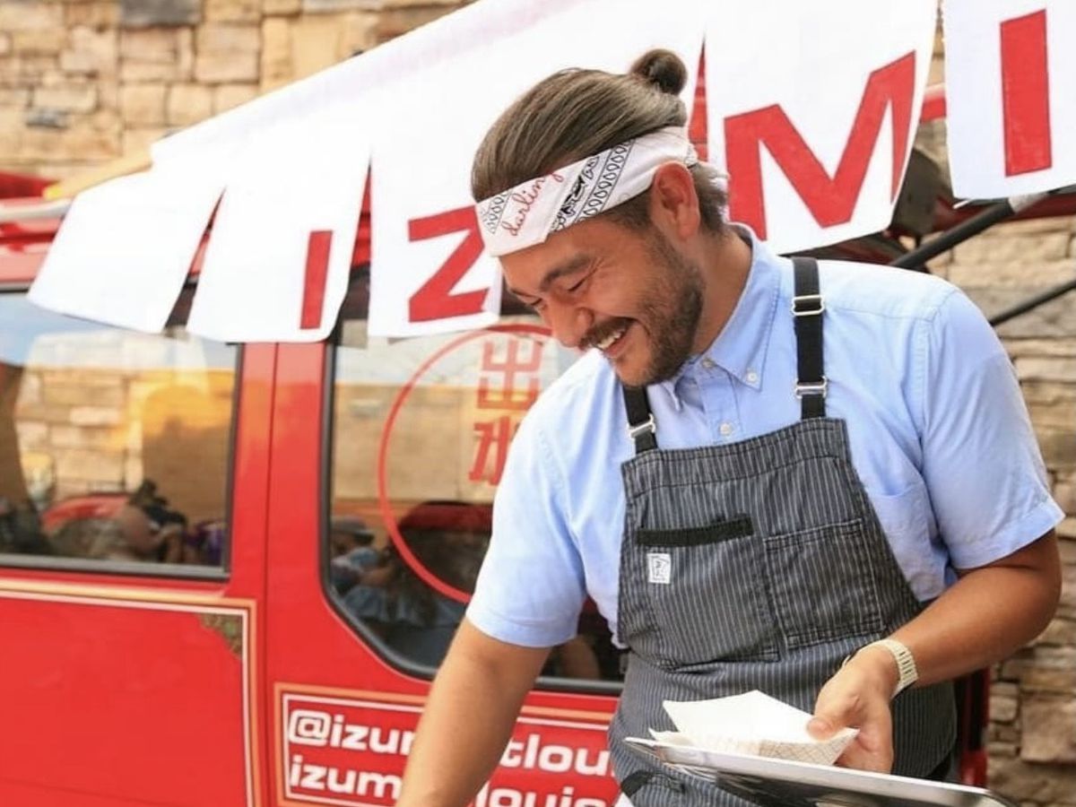 A chef in a headband stands in front of a small red firetruck and a large banner reading Izumi