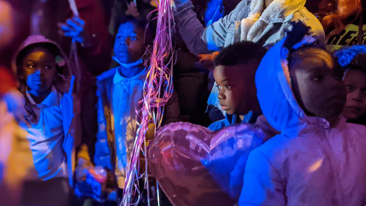 The little brother of shooting victim Phillexus Buchanan and young children stand in front of loved ones during a vigil, with blue light shining over them, and the strings of pink balloons hanging from grasped hands.