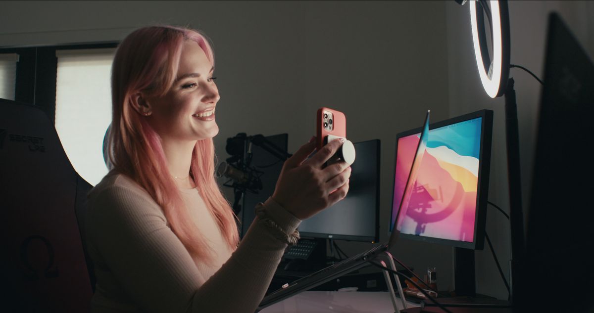 A pink-haired woman (Siri Dahl) seated at a desktop smiles while holding a cellphone camera
