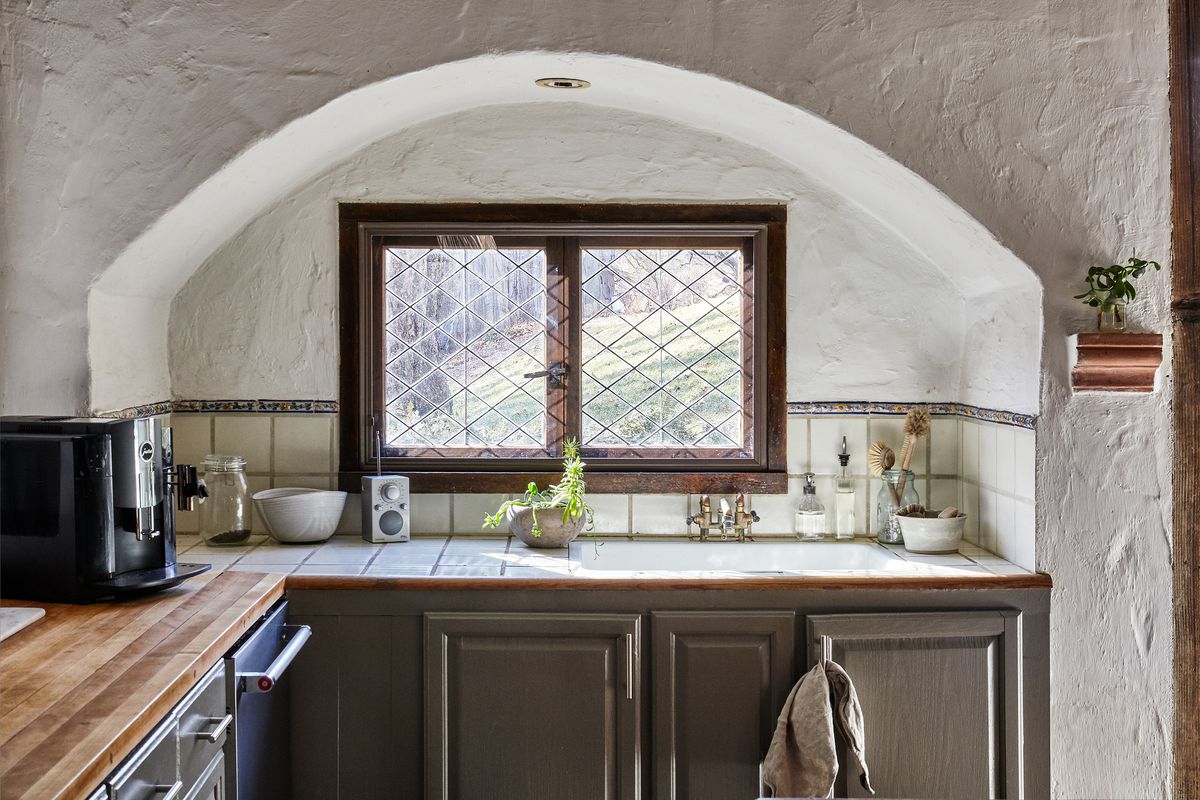 A corner of a kitchen. The walls are white, the cabinets are grey. There is an alcove cut out in the wall above the counter top with two windows.