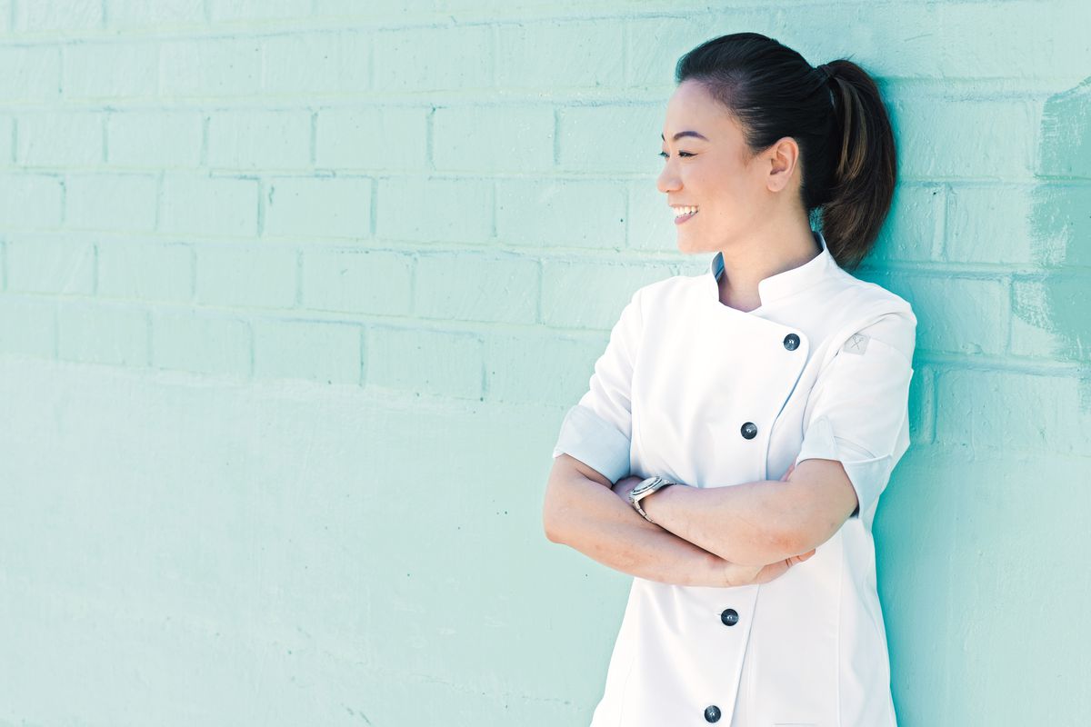 A chef in whites stands, arms crossed, next to a teal wall.