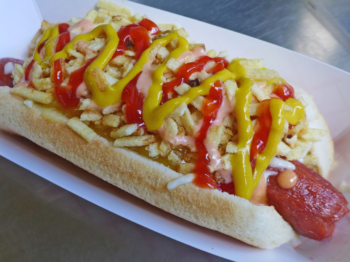 A hot dog in a bun on a metallic counter with yellow and pink sauces.