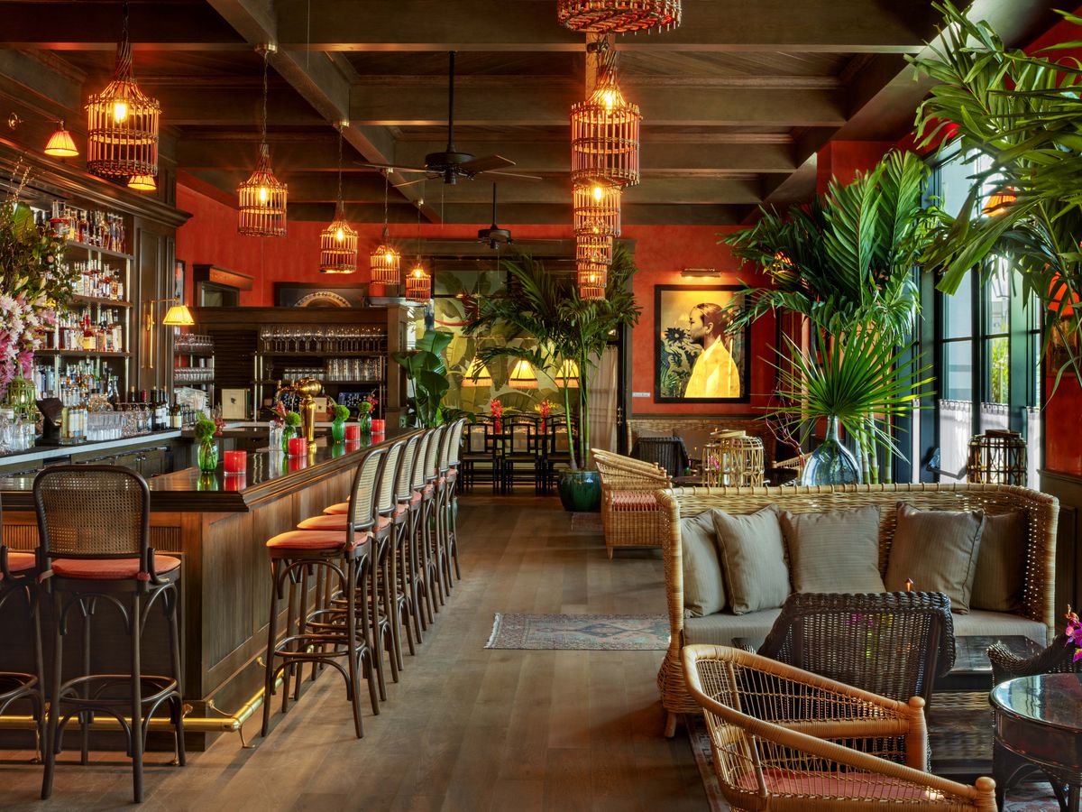 This is a photo of the inside of Le Colonial Restaurant in Delray Beach, Florida. In the foreground, on the right side are a rattan chair and a sofa. On the left side is a bar. There are palm trees and the overall look is vintage Vietnamese and tropical.