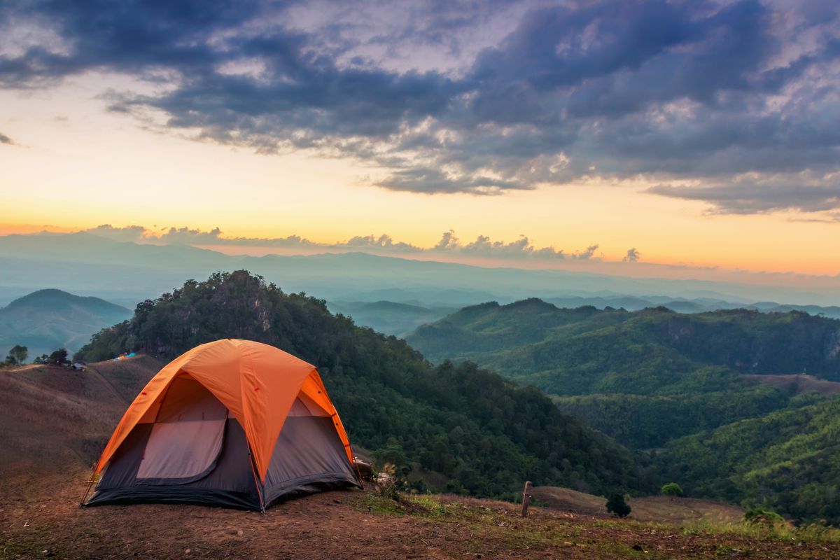 An orange tent on a mountain top. In the distance are mountains. There is a sunset and the sky is orange and purple.