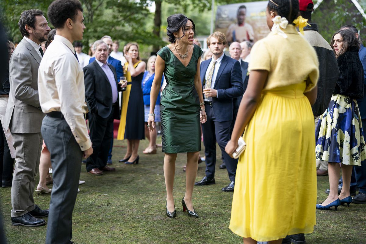 A young Black woman in a bright yellow dress stands in a crowd of people outdoors at what appears to be a garden party and yells at them in a scene from Netflix’s The Strays