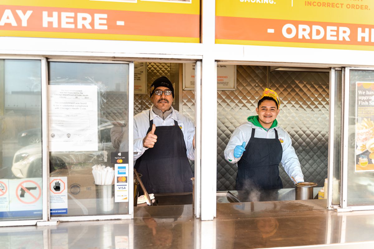 Two employees smile and pose with their thumbs up behind the counter at a street food stand.