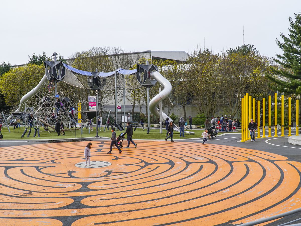 A playground with children playing. On the ground in the foreground is an orange circular design. In the distance is various assorted playground equipment. 