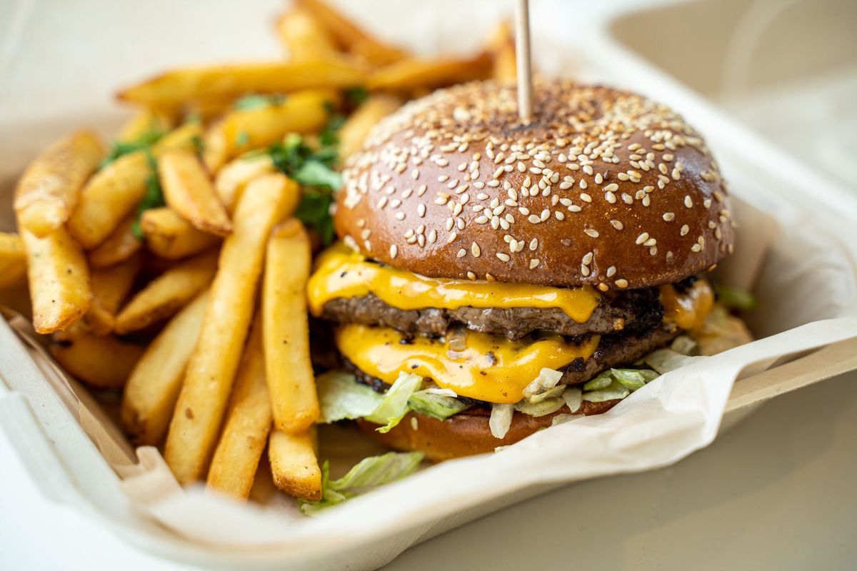 A close-up shot of a double cheeseburger with side fries in a takeout container at daytime.