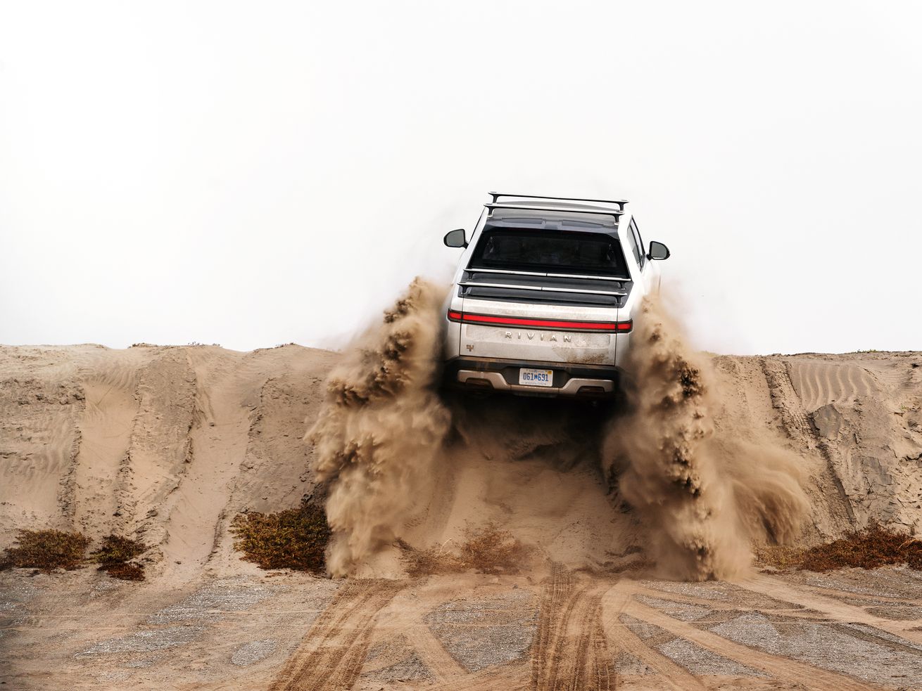 One of Rivian’s car models is seen driving away from the camera and over a pile of dirt.